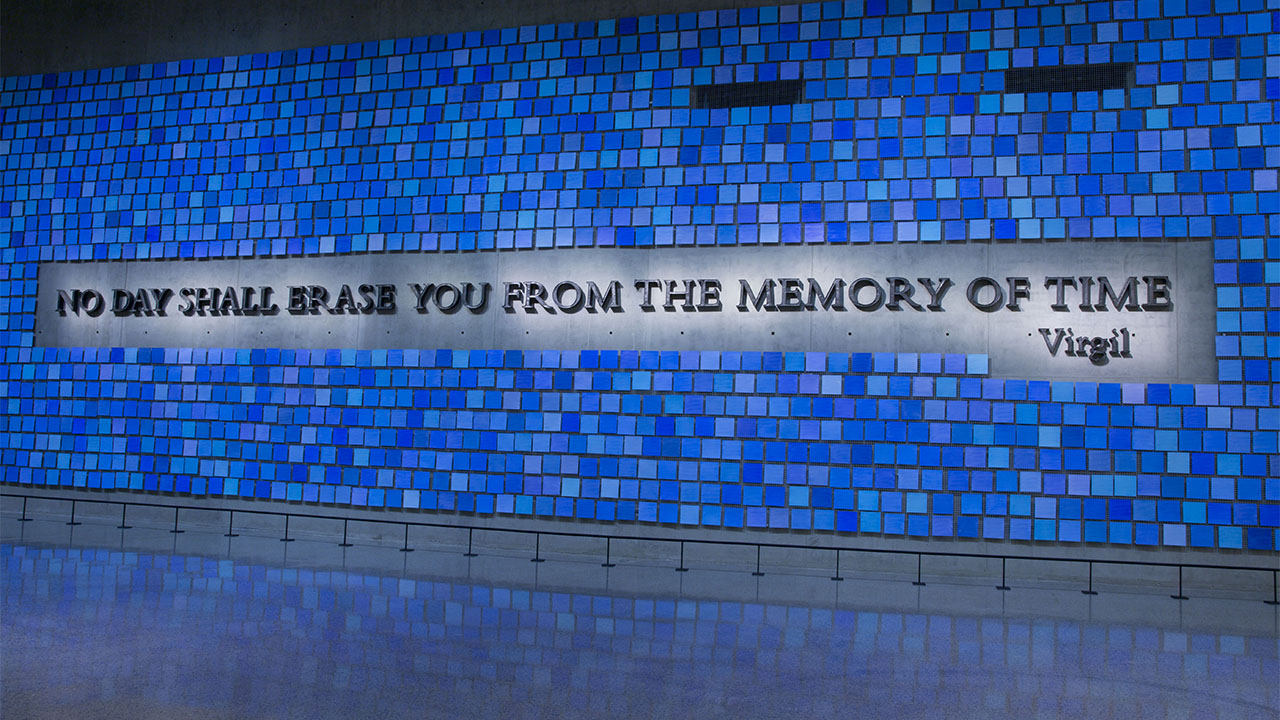 A wall of tiles, each a different shade of blue, with the inscription "No day shall erase you from the memory of time" across it. 