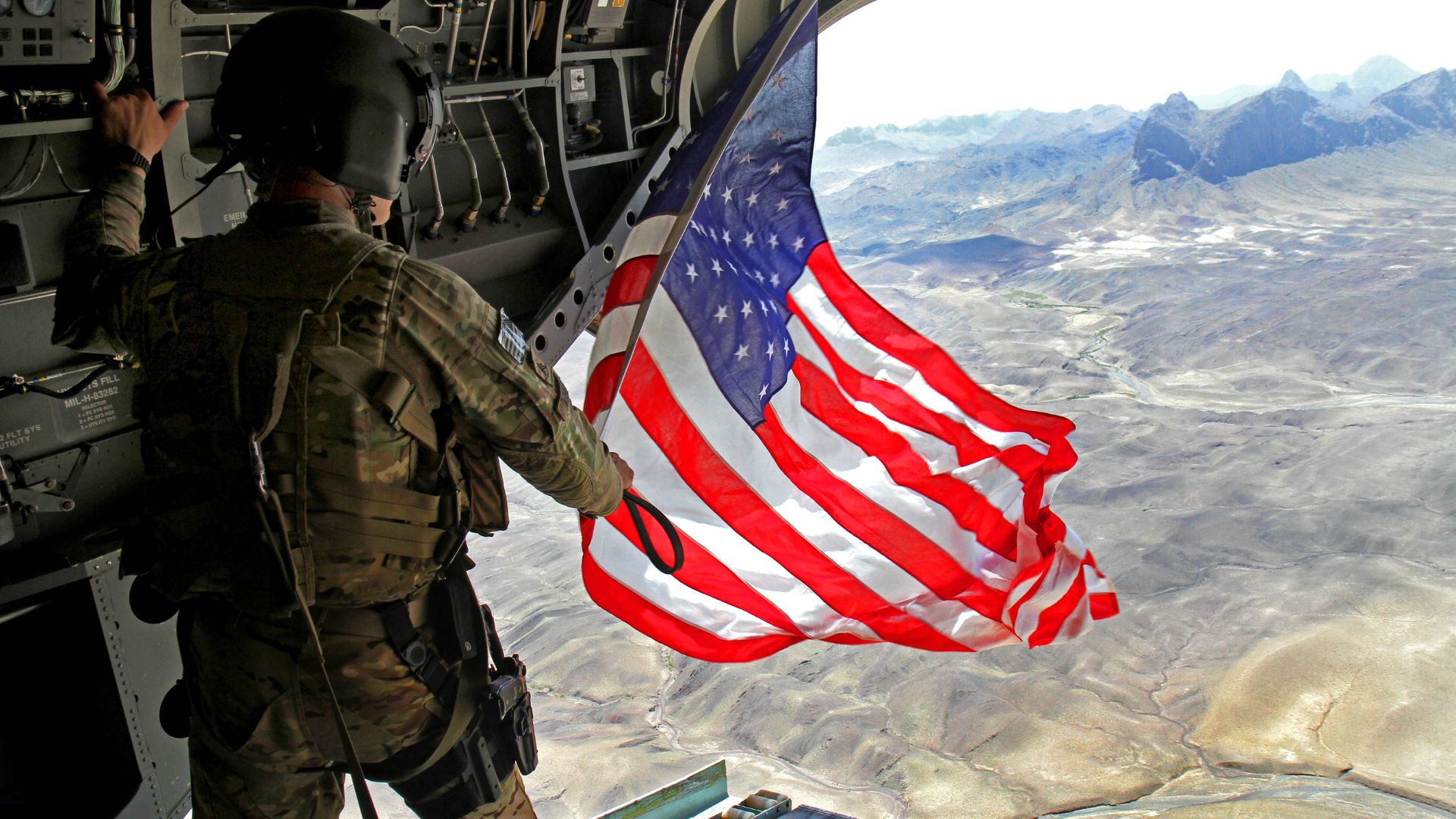 A camouflaged member of the military stands in front of an open airplane door waving an American flag. 