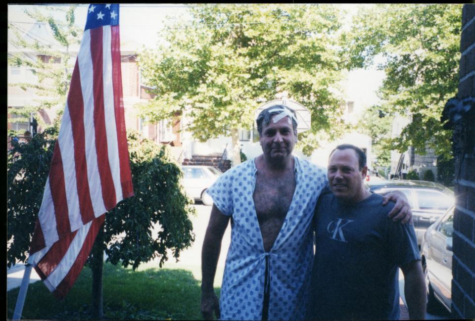 Two men, one in a hospital gown with bandages, and one in a dark shirt stand together beside an American flag