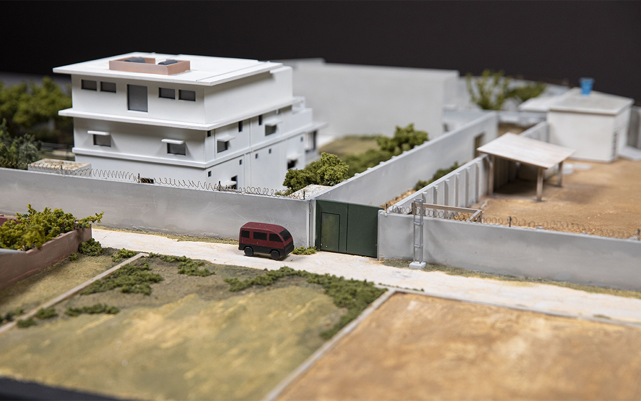 A model of the white and gray compound that housed bin Laden rests on a bed of dry-looking grass. A red courier van is parked out front.