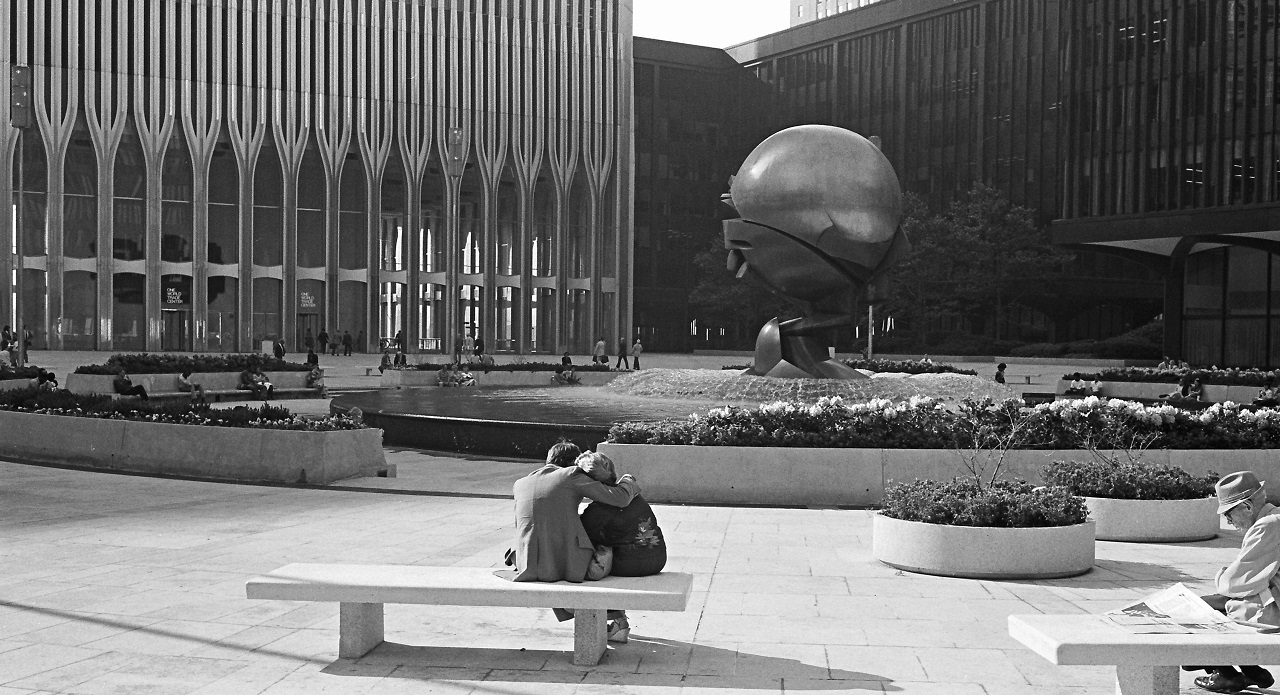 Black-and-white photo of a man sitting on a white bench with his arm around a woman, facing a spherical metal sculpture at the center. On the left is the ground level view of one of a Twin Tower.