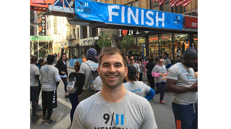 In this photograph, a man wearing a 9/11 Memorial & Museum 5k T-shirt stands underneath the finish line banner at the annual 5k Run/Walk.