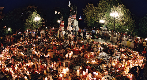 People encircling around hundreds of lit votive candles, holding vigil in a park at night.