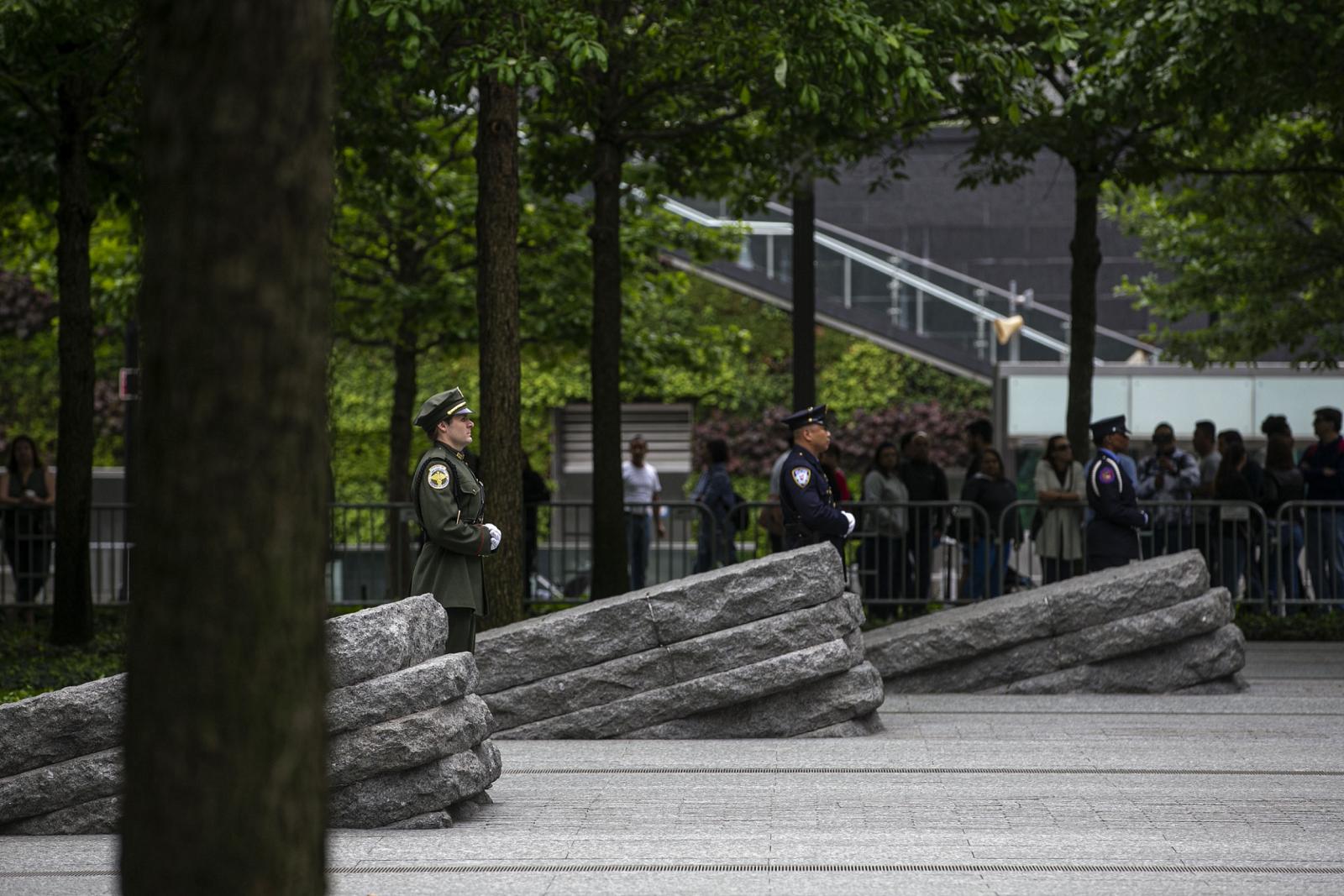 Three officials in formal uniforms stand beside three granite monoliths at the Memorial Glade. A crowd of people has gathered in the shade of trees behind them.