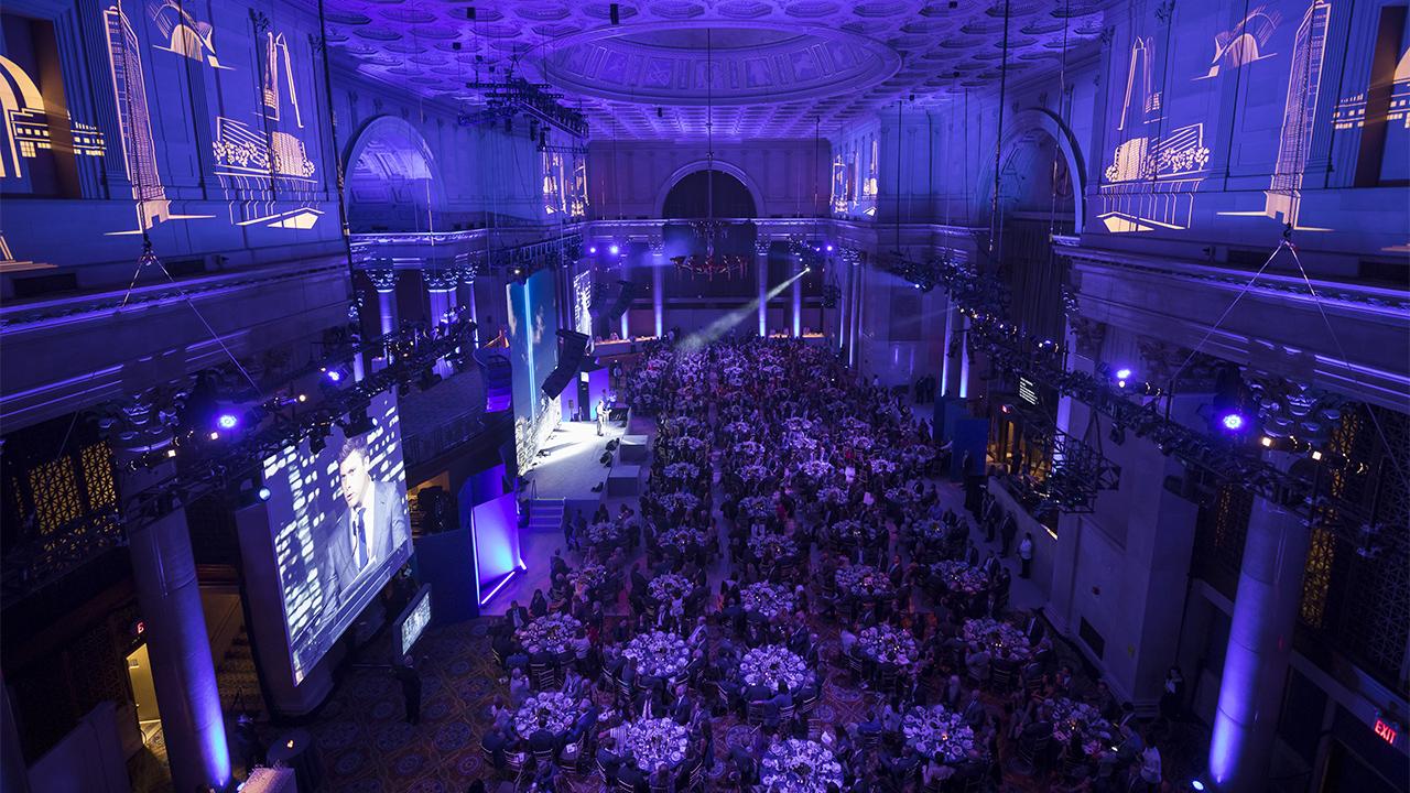 Hundreds of people sit around dozens of circular tables in a sweeping view over a large ballroom that’s lit purple. A speaker stands on stage at the center of the room with a spotlight shining on him.