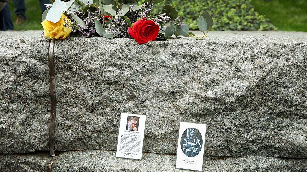 Two laminated tribute cards have been placed upright on a stone monolith at the Memorial Glade. A photo of a man is on the card to the left and a photo of a firefighter is on the card to the right. Above the cards is a bouquet of flowers, including a yellow rose and a red rose.