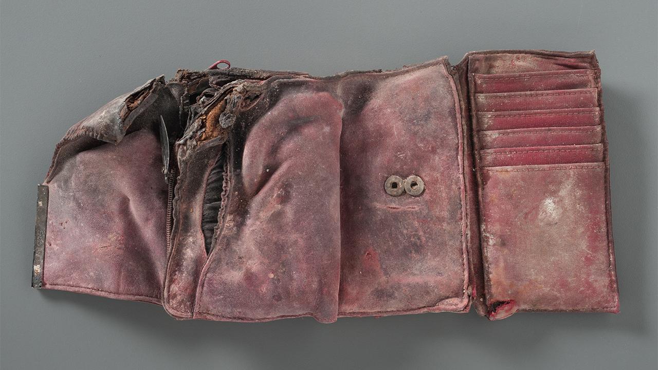 This recovered red wallet, belonging to Giovanna Galletta Gambale, shows significant damaged with encrusted dust and warped from exposure to the elements. There are scorch marks and soot stains on the fabric.