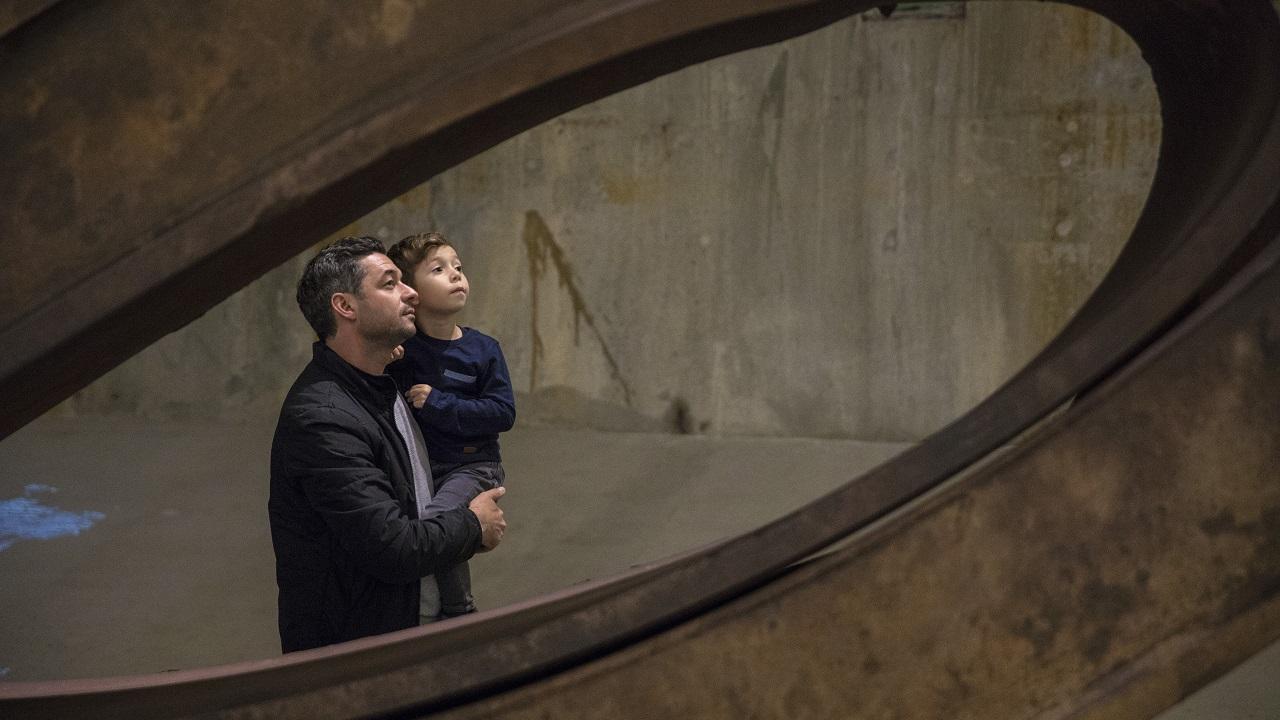 A man holds a boy as the two observe an object out of view in Foundation Hall. The two of them are framed by a piece of bent structural beam from the World Trade Center.