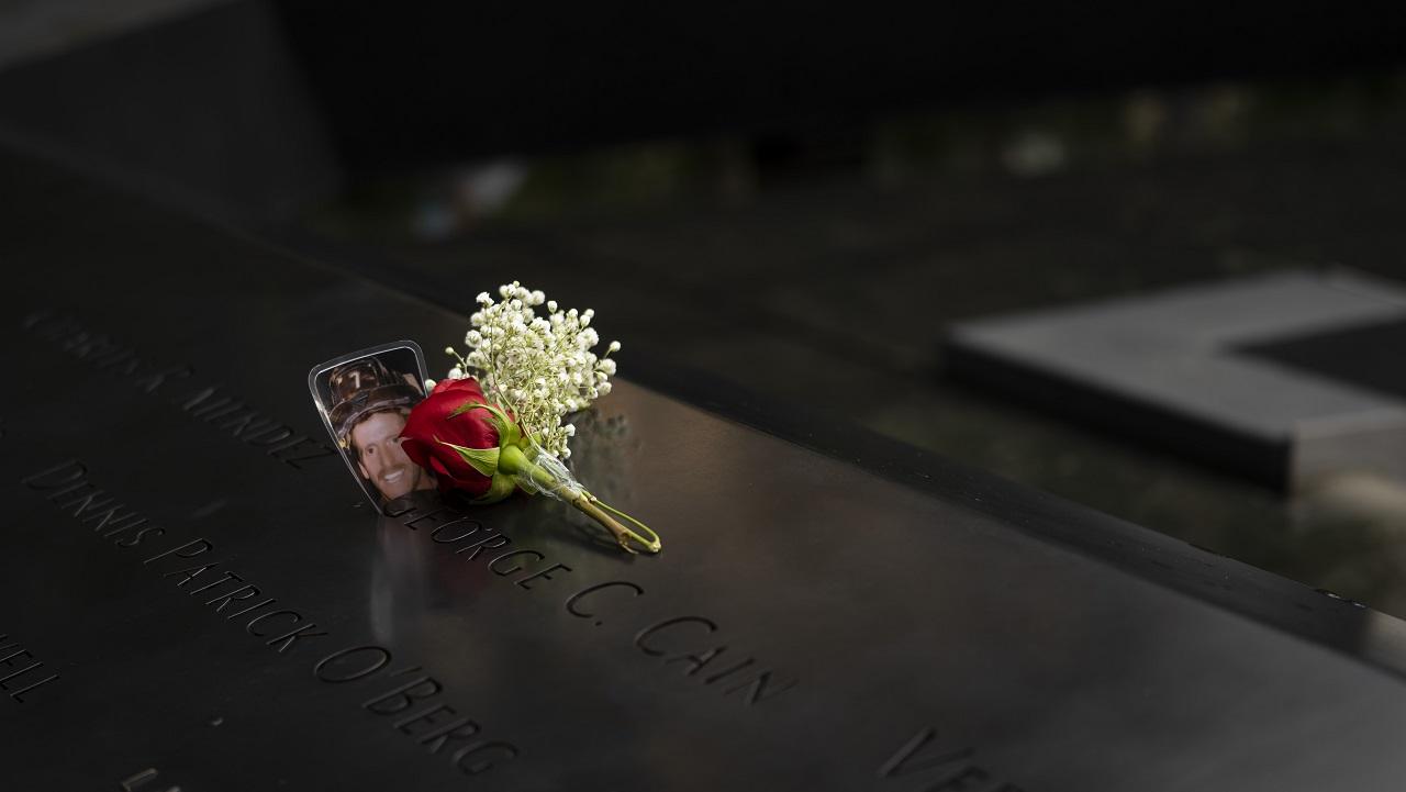 A laminated photo of a firefighter wearing a helmet rests inside a name on the bronze parapet of the Memorial alongside single red rose, surrounded by small white flowers.