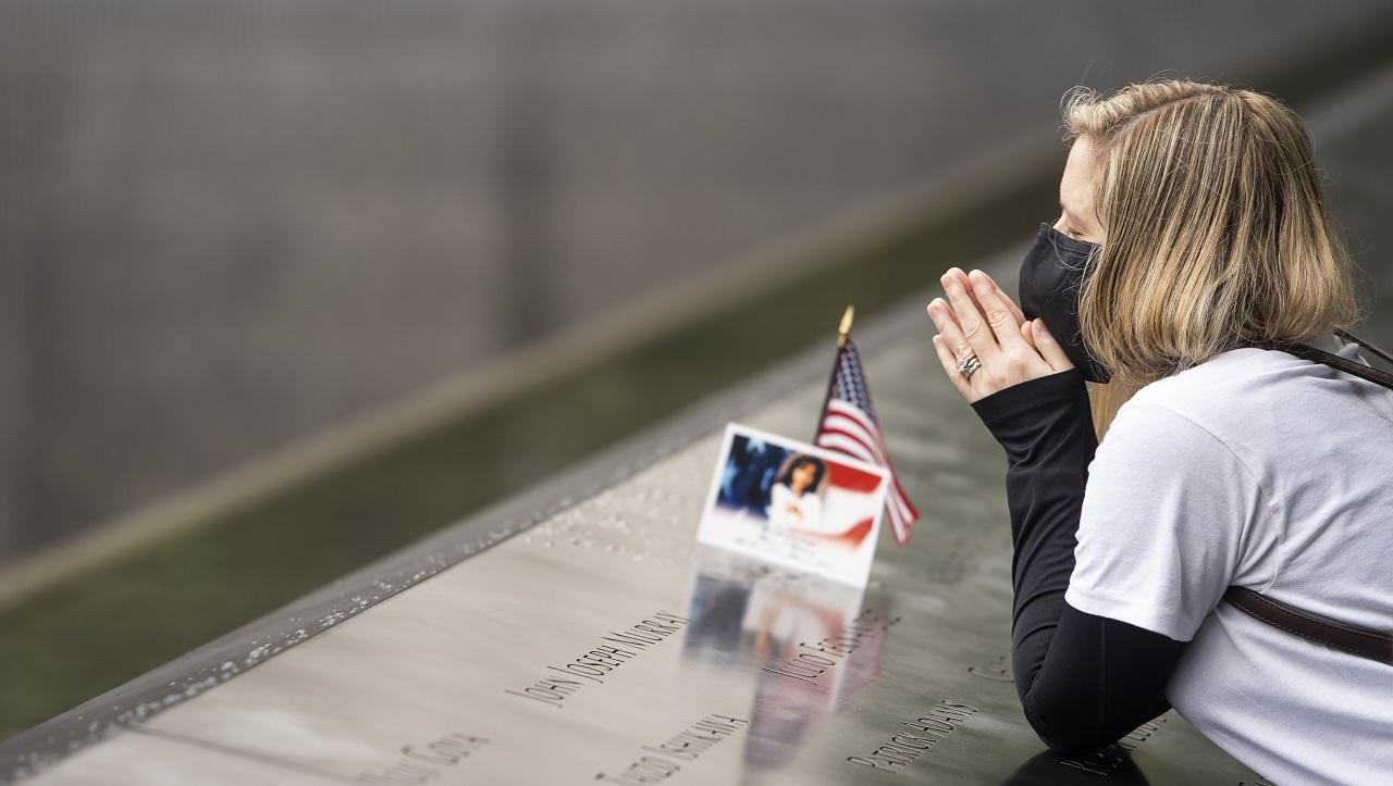 A women holds her hands, clasped in prayer, near her face as she leans on the 9/11 Memorial. A name on the Memorial is adorned with a photograph and American flag as this visitor closes her eyes in remembrance and prayer.