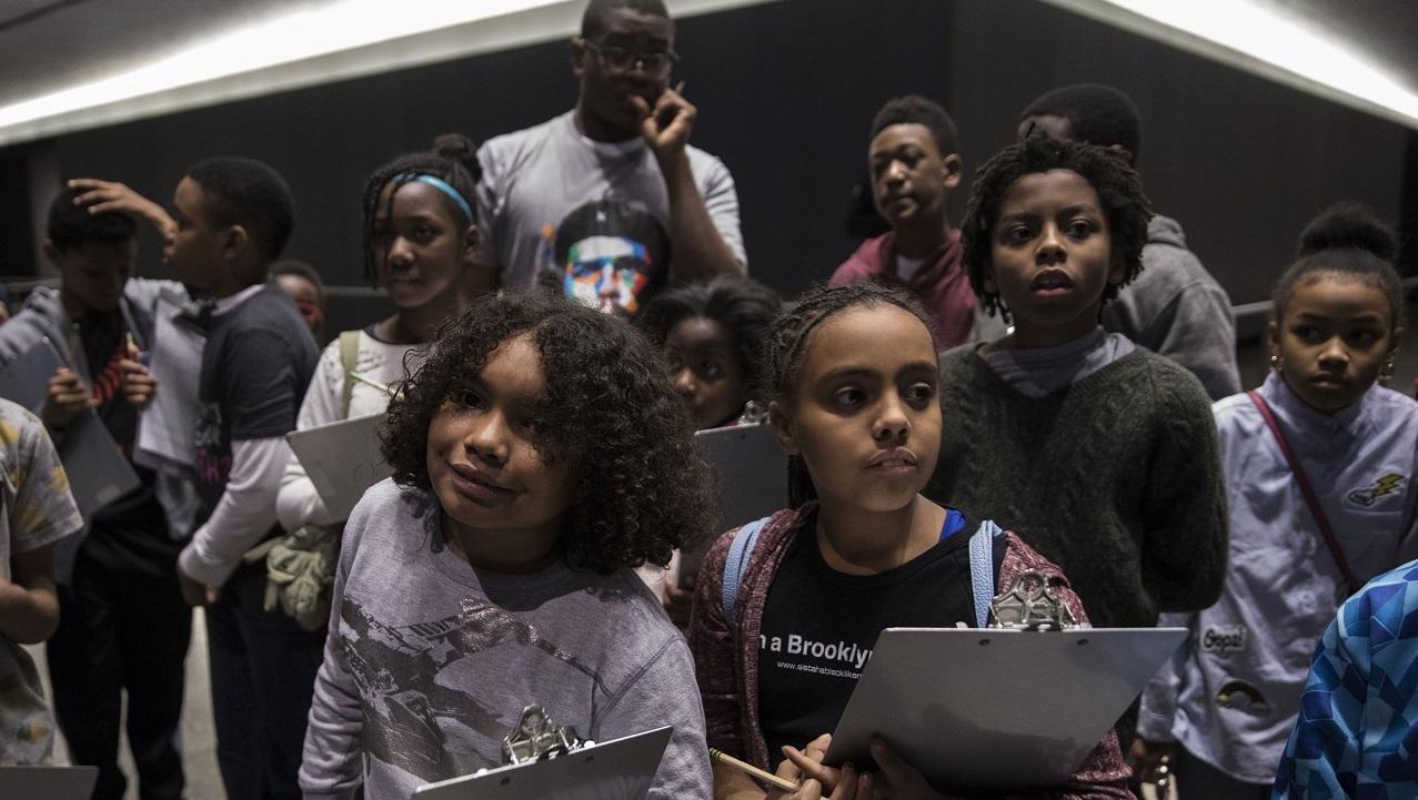 A grouping of elementary school students holding clipboard smile softly on an educational tour of the 9/11 Memorial Museum.