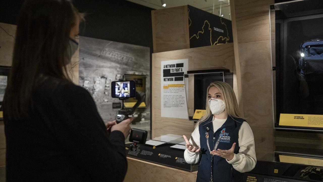 A tour guide wearing a mask stands in the "Revealed" exhibition space to give a virtual tour. Another Museum staff member films the tour with her back to the camera.
