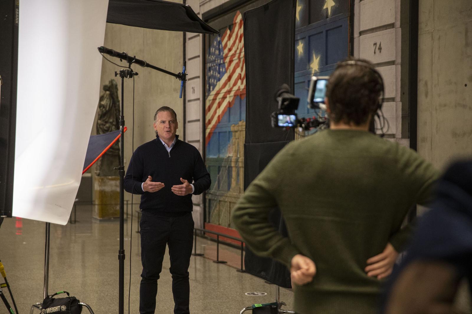 A man in a dark sweater speaks in front of a camera. 