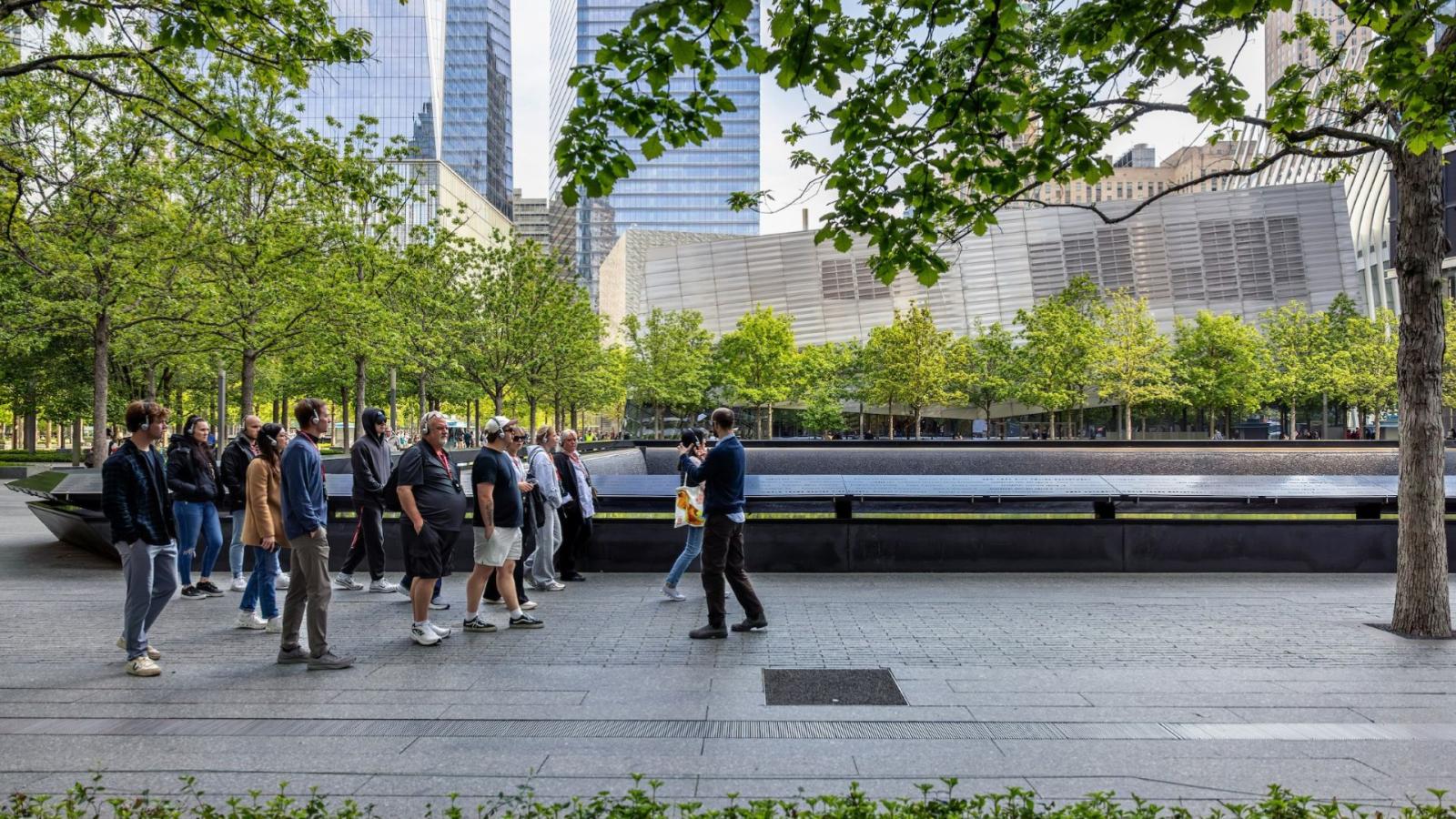 A group with a guide stands on the Memorial plaza with one of the reflecting pools to their left