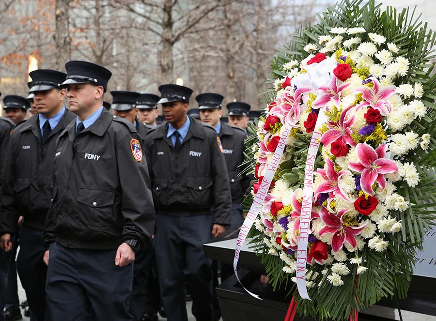 About a dozen probationary FDNY firefighters are formally dressed as they walk by a brightly colored wreath at Memorial plaza. The wreath reads “In memory of those who made the supreme sacrifice FDNY.”