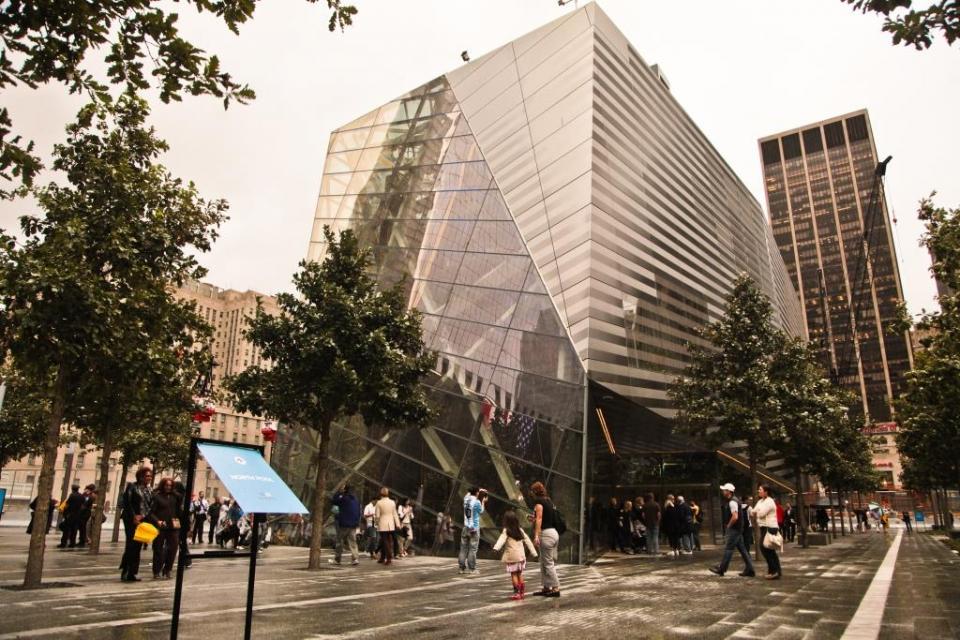 Dozens of visitors walk around the glass and steel facade of the Museum pavilion on Memorial plaza.