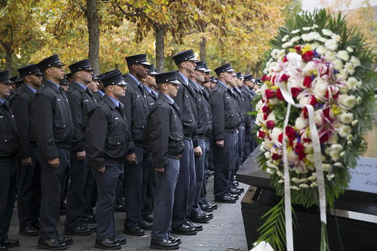 Probationary firefighters line up along the south reflecting pool near the names of FDNY victims. A large wreath stands in the foreground.