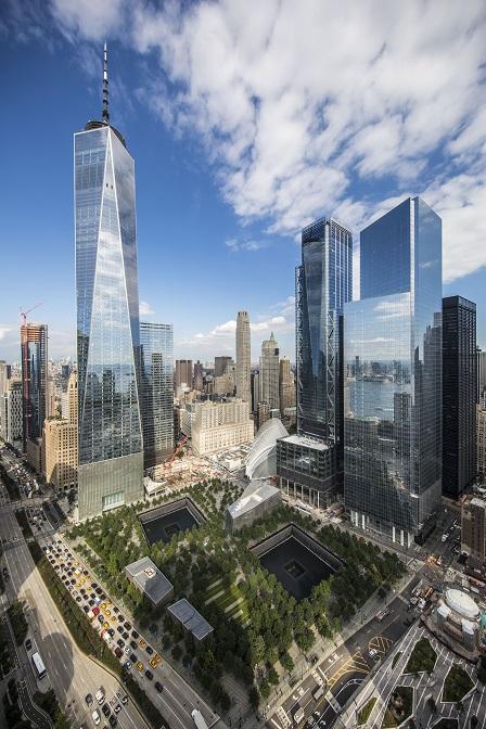 An aerial view shows One World Trade Center and the 9/11 Memorial on a partly cloudy day. The buildings of lower Manhattan stand in the distance.