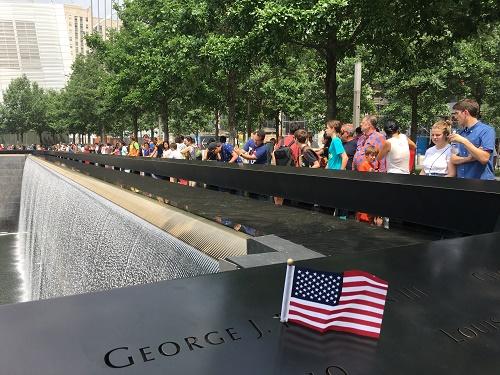 Visitors observe a reflecting pool at the Memorial. A small American flag has been placed at a name on a bronze parapet in the foreground.