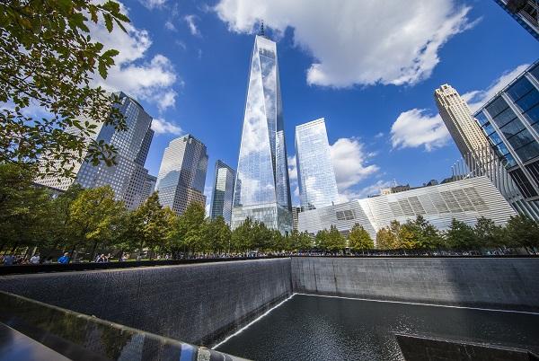 One World Trade Center and surrounding skyscrapers tower over Memorial Plaza. Clouds in a blue sky cast a shadow on the Memorial below.