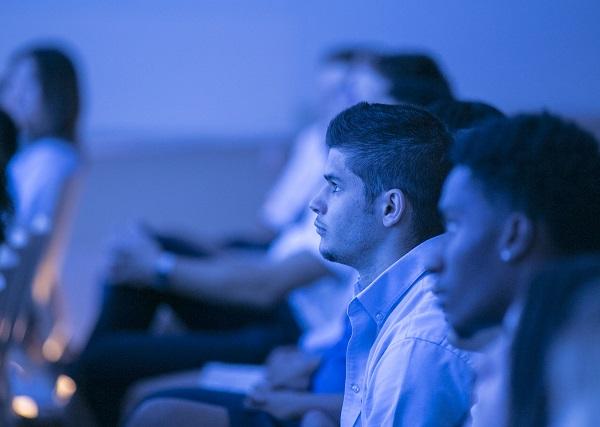 Blue light from the Museum auditorium shines on a student as he looks towards the stage during an event. Other students watch beside him.