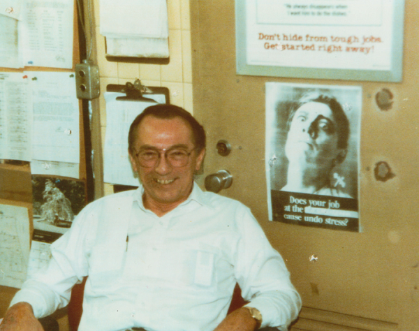 This historical photo shows Robert Kirkpatrick in a white collared shirt in his office at the World Trade Center. Papers and images are tacked onto the wall behind him. 