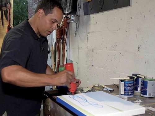 Frederico A. Ruiz works on a piece of art in his studio. He is applying paint from a red bottle onto a small canvas.