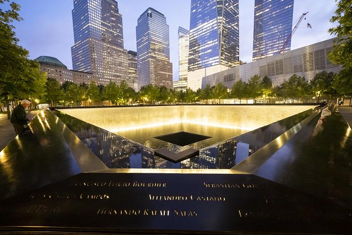 The rim of the 9/11 Memorial pool is illuminated from inside. A man leans over the side of the names parapet. Skyscrapers surround the scene at dusk.