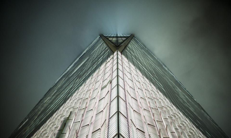 A view of One World Trade Center from street level shows the skyscraper rising into the clouds on an overcast night. Only part of the building is visible below the clouds.