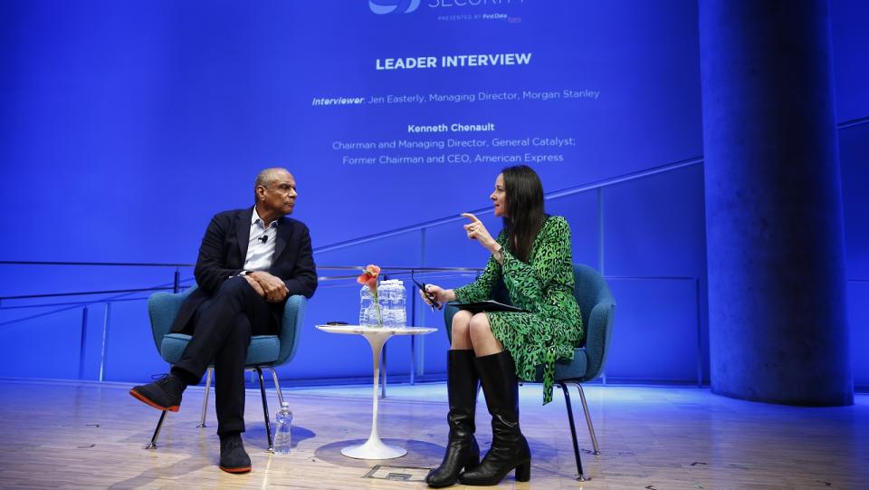 A man and a woman are seated on a stage in the auditorium of the 9/11 Memorial Museum discussing security issues before a blue display at the back of the stage..  