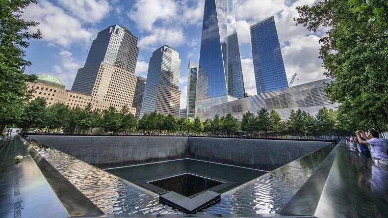 A photo taken at the southwest corner of the 9/11 Memorial plaza shows One World Trade Center emerging through the trees. Clouds an blue sky is reflected in the building's facade.