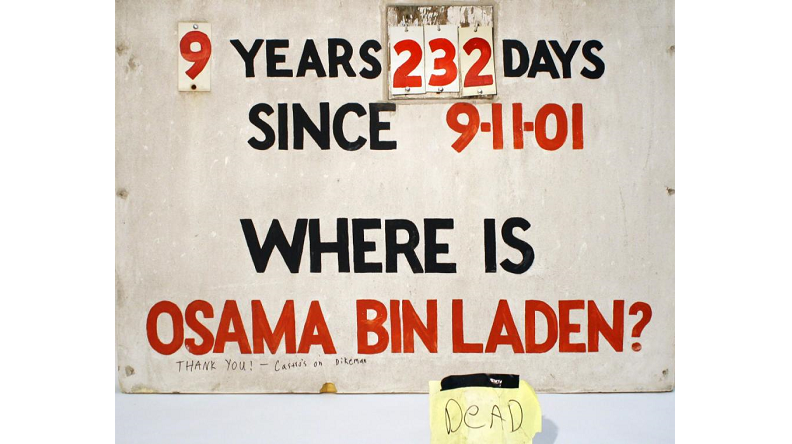 A sign that tracked the days Osama bin Laden was at large is displayed on a white surface. The sign reads: “9 years, 232 days since 9/11/01. Where is Osama bin Laden?” A yellow piece of paper beside the sign reads “Dead.”